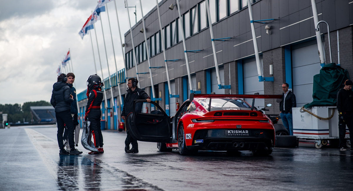Ein Team steht in Assen um den roten Porsche-Rennwagen in der nassen Boxengasse vor wehenden Flaggen