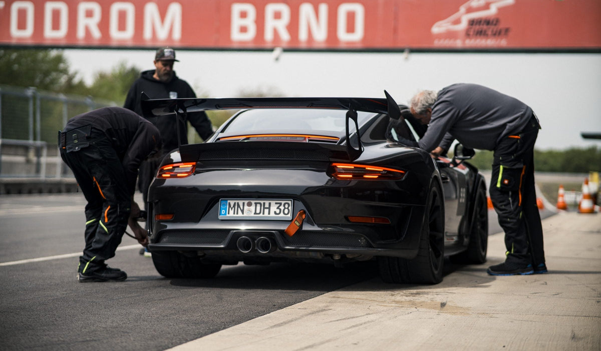 Drei Boxencrew-Mitglieder beugen sich über einen dunklen Porsche vor dem Schriftzug Automotodrom Brno