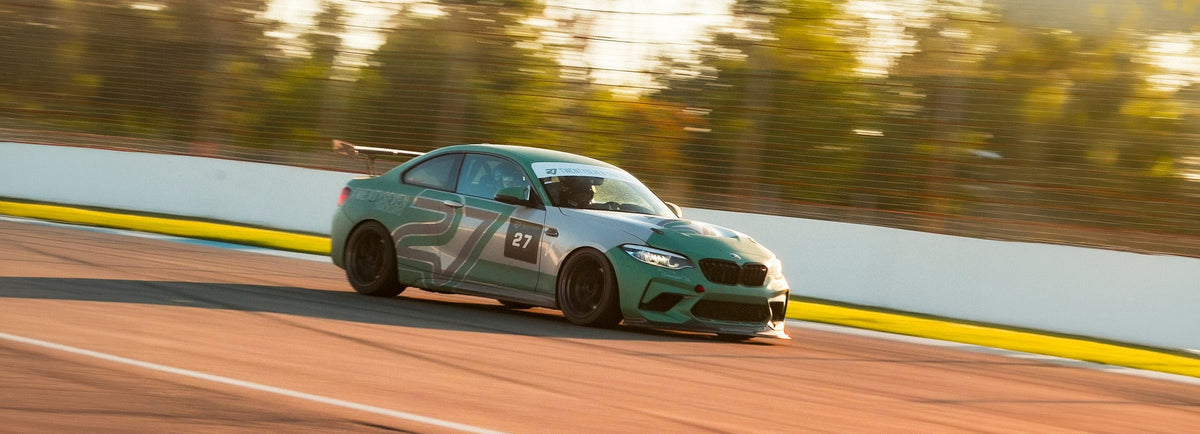 24/7-Performance-BMW M2 Competition in grün-grauer TWENTYSEVEN-Livery mit Startnummer 27, Frontansicht im Gegenlicht der Abendsonne auf der Rennstrecke