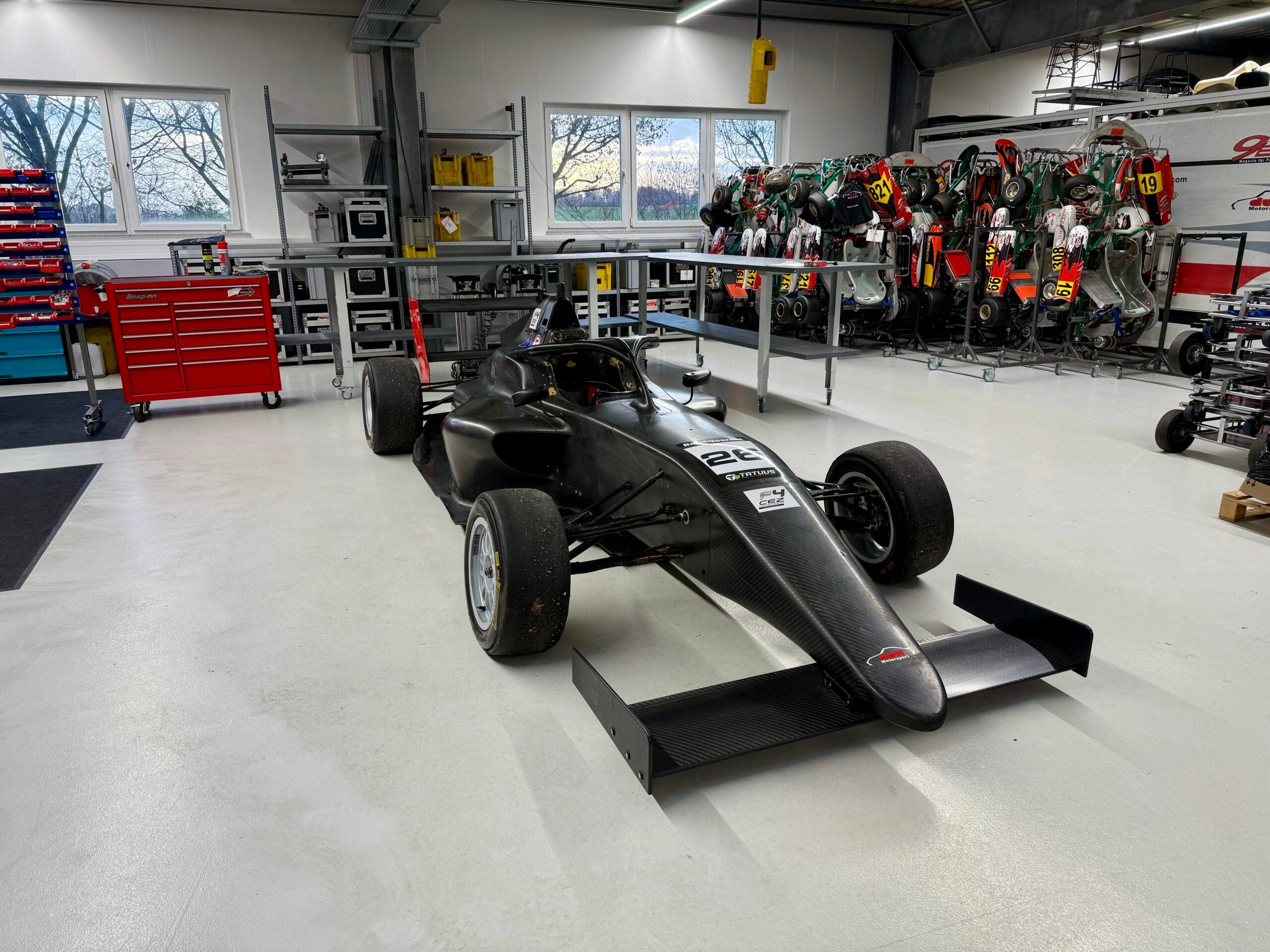 Schwarzes Formelfahrzeug in einer Motorsport-Werkstatthalle mit Karts und Werkzeug im Hintergrund.