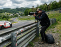 Videograf am Rand der Nürburgring Nordschleife. Auf der Rennstrecke fährt ein Porsche 992 GT3R mit dem Porsche Motorsport Livery vorbei.