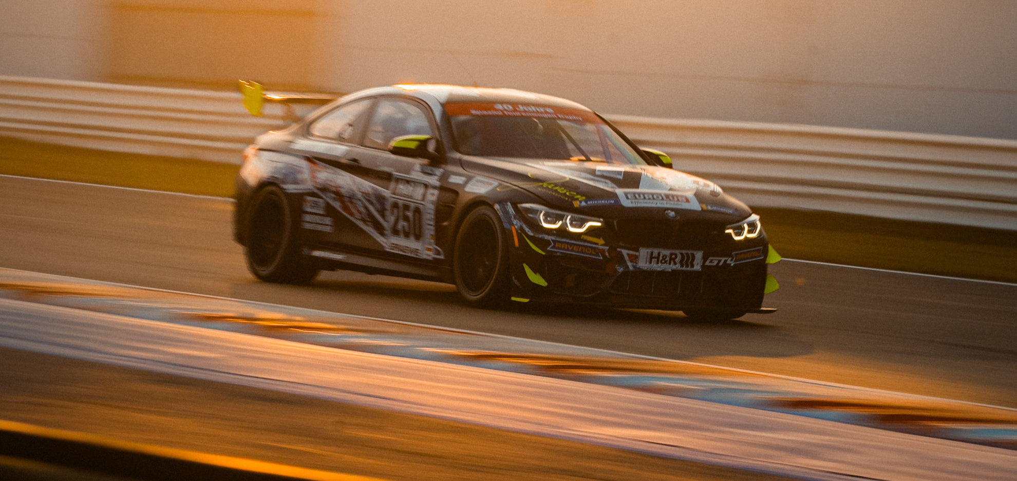 BMW F82 M4 GT4 in der BMW Motorsport Junior Lackierung auf einer Rennstrecke während eines Sonnenuntergangs.