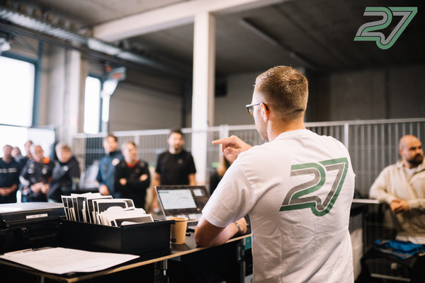 Ein Sprecher im weißen Shirt deutet auf eine Gruppe Zuhörender beim Fahrerbriefing in der Halle