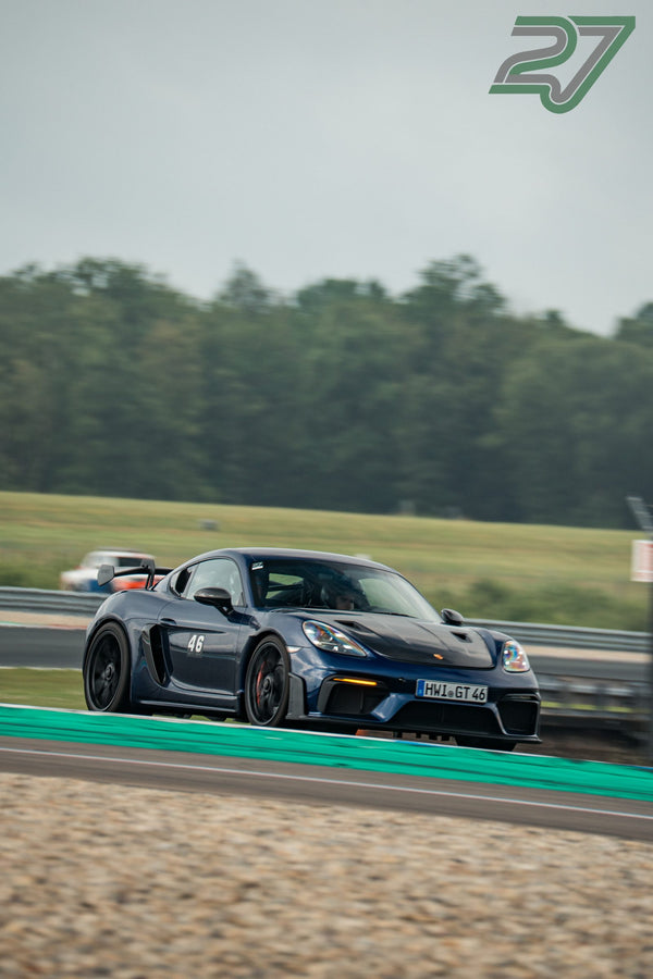Dunkelblauer Porsche 718 Cayman GT4 fährt in Assen auf der sonnigen Geraden vor dichtem Wald
