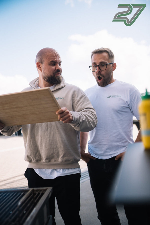 Zwei Männer schauen überrascht auf eine Holzplatte, die ihnen in die Kamera gehalten wird