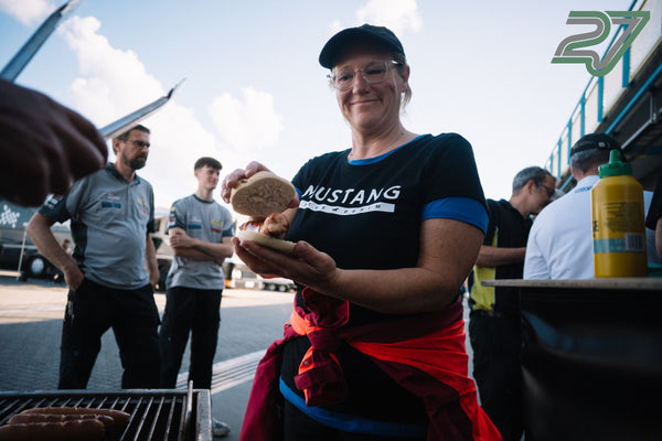 Lächelnde Frau mit Cap hält ein frisch belegtes Hot-Dog-Brötchen, im Vordergrund brutzeln Würstchen