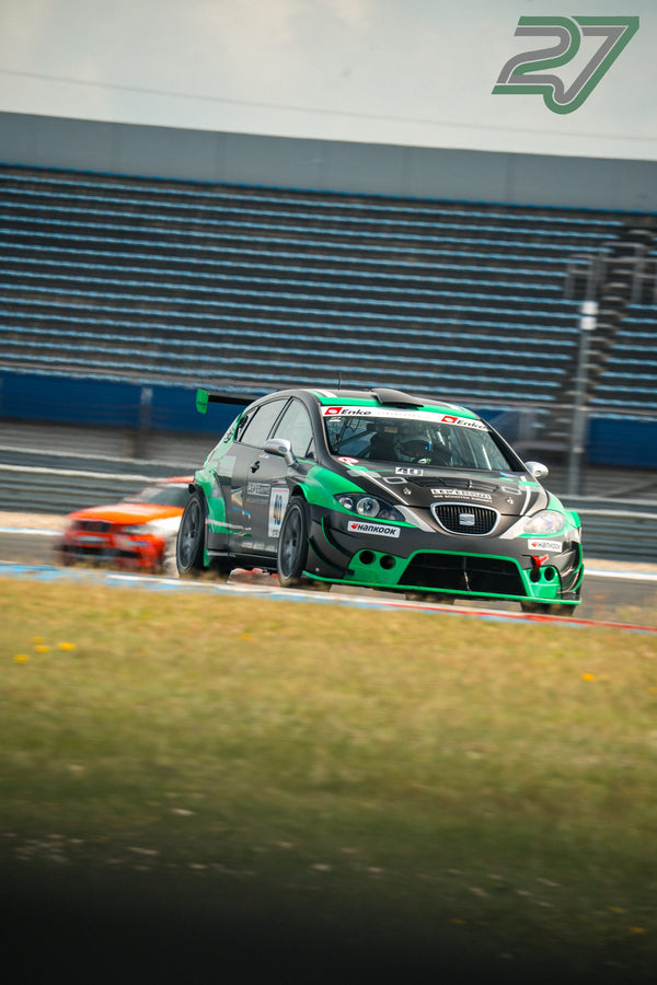 Grün-schwarzer SEAT Leon TCR führt unter bedecktem Himmel vor einem verschwommenen roten Fahrzeug