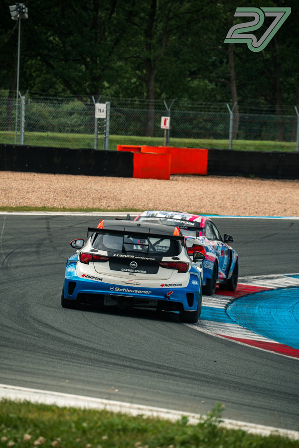 Blau-weißer Opel Astra TCR führt von hinten gesehen vor einem pink lackierten Verfolger durch schattigen Wald