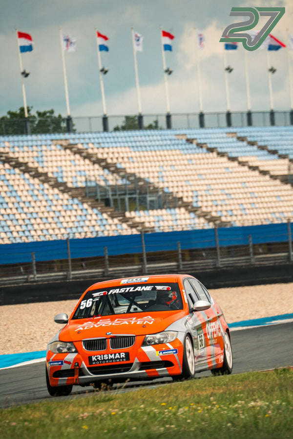 Orangefarbener BMW-Rennwagen kommt schräg von vorne heran, Flaggen wehen über der leeren Tribüne in Assen