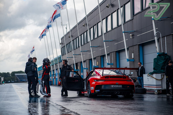 Ein Team steht in Assen um den roten Porsche-Rennwagen in der nassen Boxengasse vor wehenden Flaggen