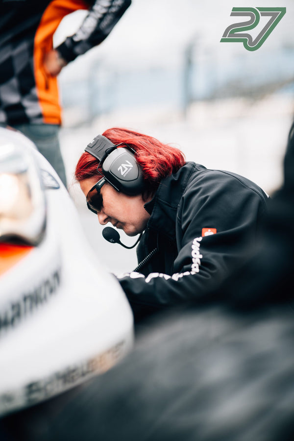Eine rothaarige Mechanikerin mit Headset und Sonnenbrille beugt sich konzentriert über den Rennwagen
