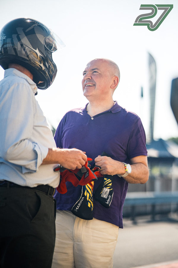 Ein Fahrer mit Helm übergibt lächelnd rote Rennhandschuhe an einen Mann im violetten Polohemd