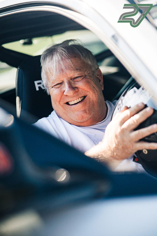 Ein älterer Fahrer lacht breit im Cockpit und hält ein Blatt Papier in der erhobenen Hand