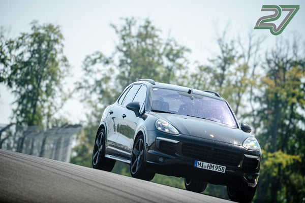 Schwarzer Porsche Cayenne kommt über eine Kuppe, Vorderräder heben leicht ab, Bäume im Hintergrund