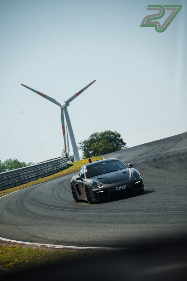 Mattgrauer Porsche Cayman zieht eine Kurve, im Hintergrund dreht sich ein Windrad am Horizont