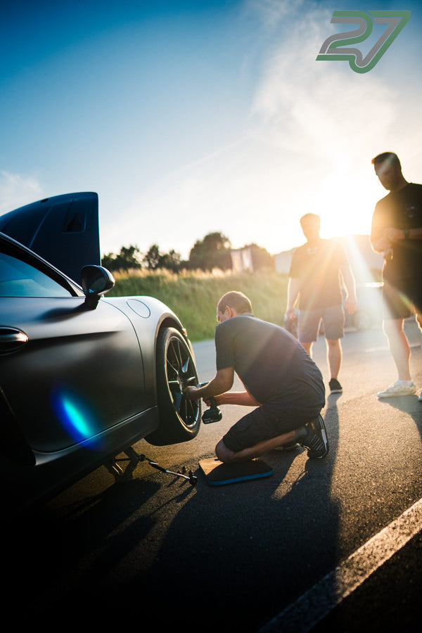Ein Mechaniker wechselt im Gegenlicht das Rad an einem dunklen Porsche mit GTS-Schriftzug, zwei Männer schauen zu