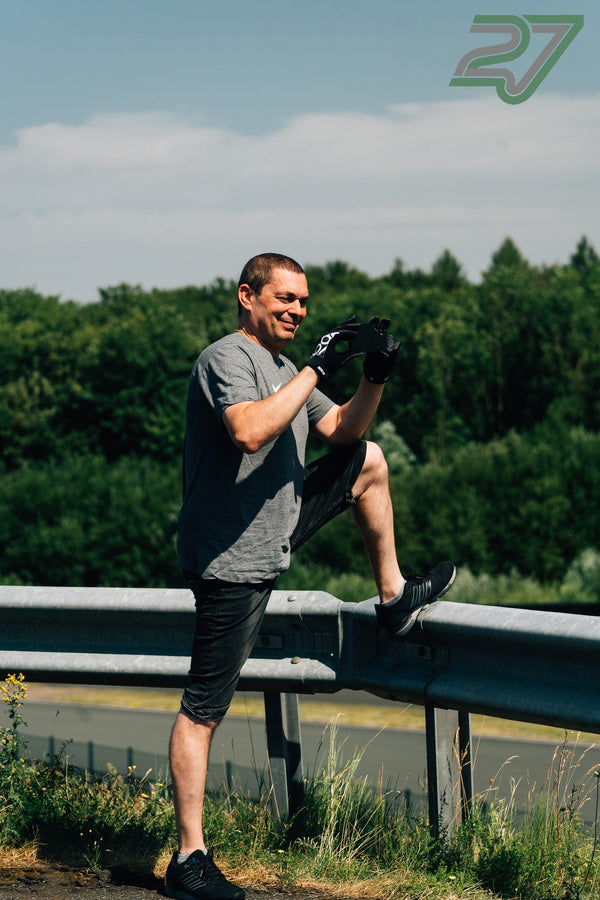 Ein Mann mit Rennhandschuhen filmt lächelnd mit dem Handy, ein Bein auf der Leitplanke abgestützt