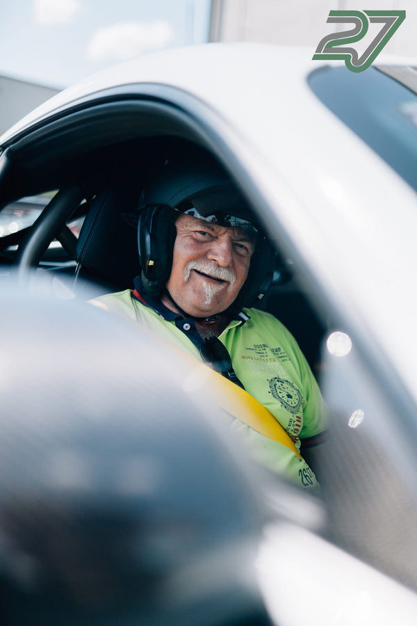 Ein Fahrer mit Headset und dunklem Visier lächelt breit hinter dem Lenkrad, der Sicherheitsgurt ist angelegt
