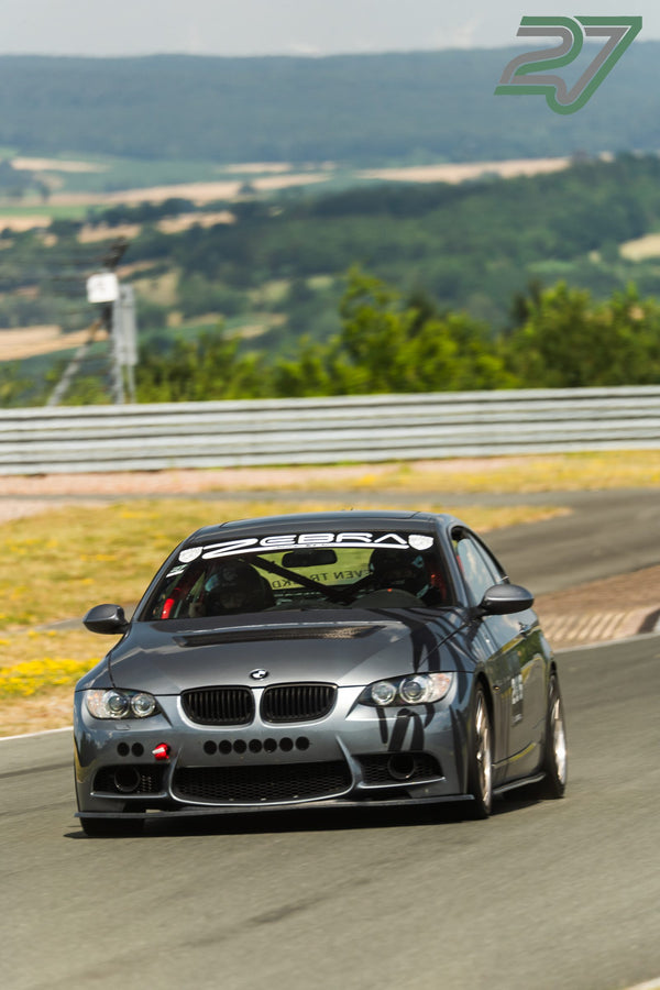 Ein anthrazitfarbener BMW M3 mit Zebra-Racing-Aufkleber fährt auf die Kamera zu, weite Hügellandschaft im Hintergrund