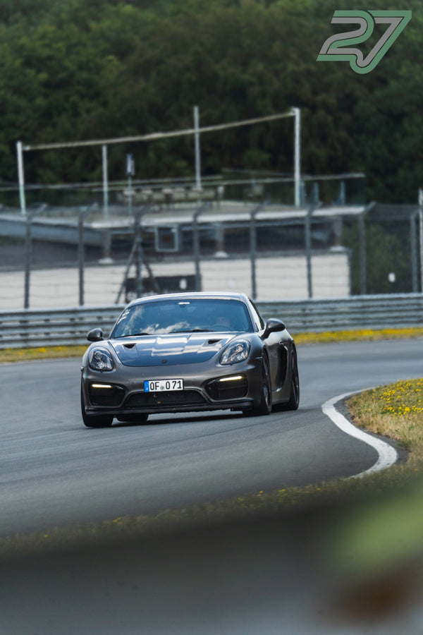 Mattgrauer Porsche Cayman fährt im hellen Sonnenlicht frontal durch eine Kurve auf die Kamera zu