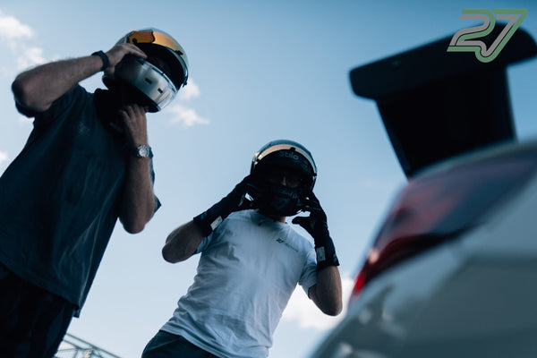 Zwei Männer setzen im Gegenlicht ihre Helme auf, daneben steht ein Sportwagen mit geöffneter Heckklappe