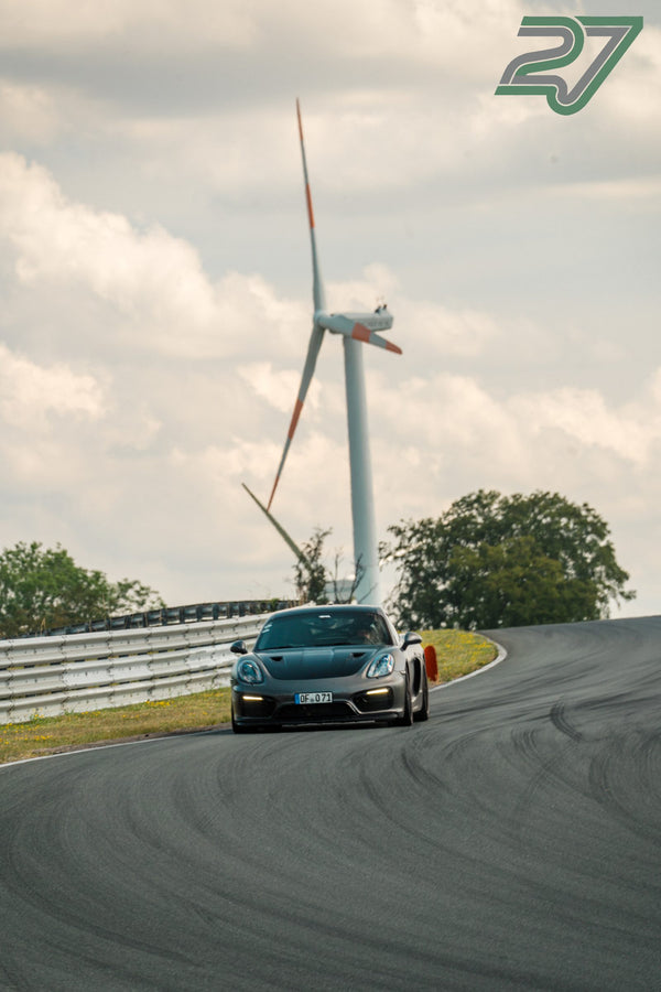 Ein dunkler Sportwagen fährt auf eine Kurve zu, im Hintergrund dreht sich ein großes Windrad am Bilster Berg