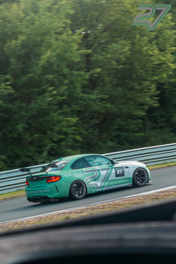 Ein mintgrüner BMW M2 mit Teambeschriftung und großem Heckflügel fährt dicht am Waldrand entlang