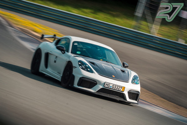 Ein weißer Porsche Cayman GT4 RS neigt sich in einer schnellen Kurve, die Leitplanke verschwimmt im Hintergrund