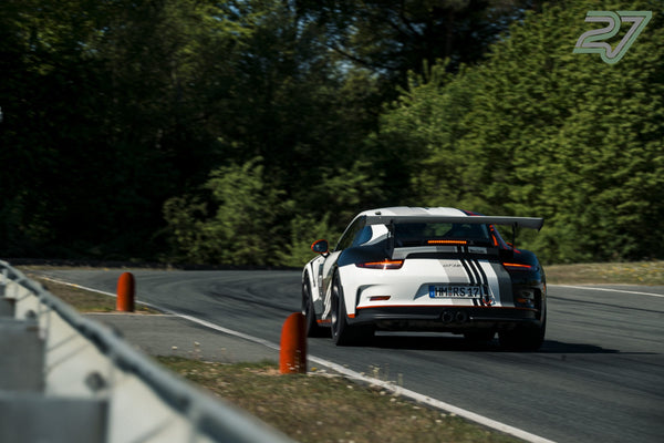 Weißer Porsche mit GT3-RS-Streifen und großem Heckflügel fährt aus der Waldkurve heraus