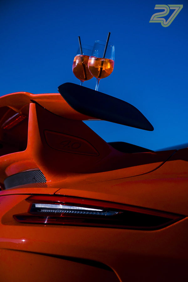 Zwei Cocktailgläser balancieren auf dem Heckflügel eines orangen Porsches vor blauem Abendhimmel