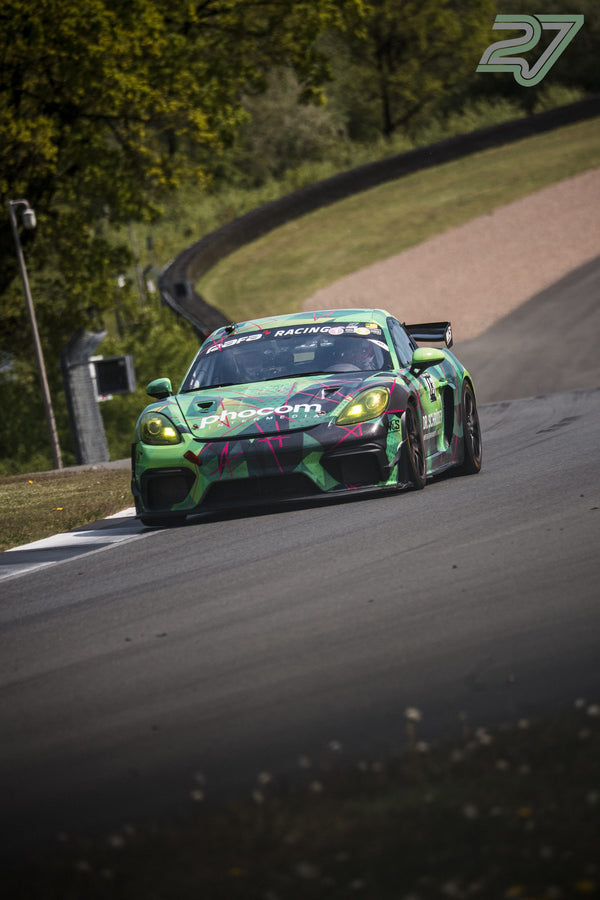 Grün-pink foliierter Porsche Cayman-Rennwagen kurvt vorbei an Kiesbett und Leitplanke am Bilster Berg