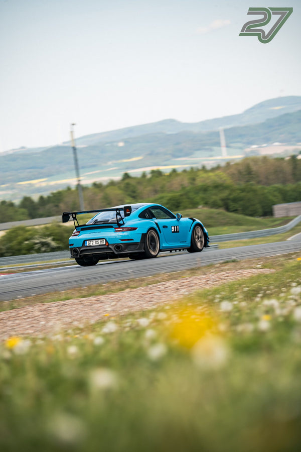 Hellblauer Porsche 911 mit Heckflügel neigt sich in die Kurve vor der Hügellandschaft am Bilster Berg