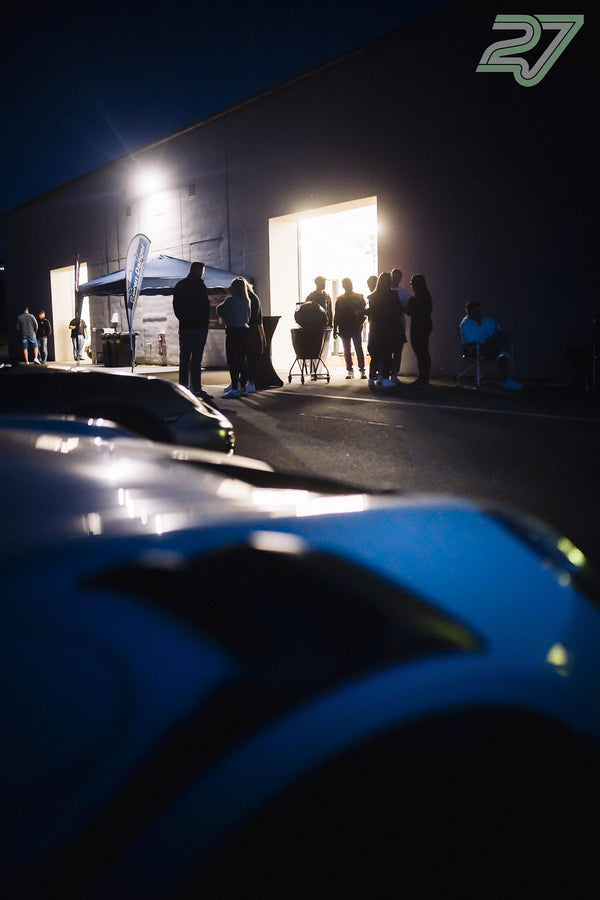 Ein blaues Auto steht im Vordergrund, während Teilnehmer vor der beleuchteten Halle plaudern