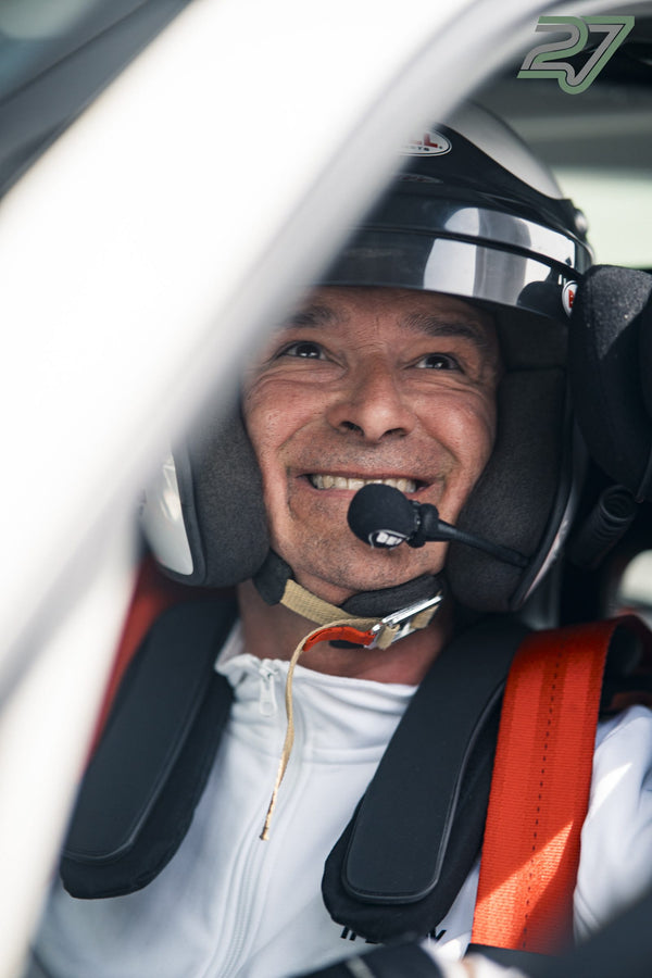 Ein Fahrer mit Helm und Kopfhörer-Mikrofon lacht breit angeschnallt im Cockpit