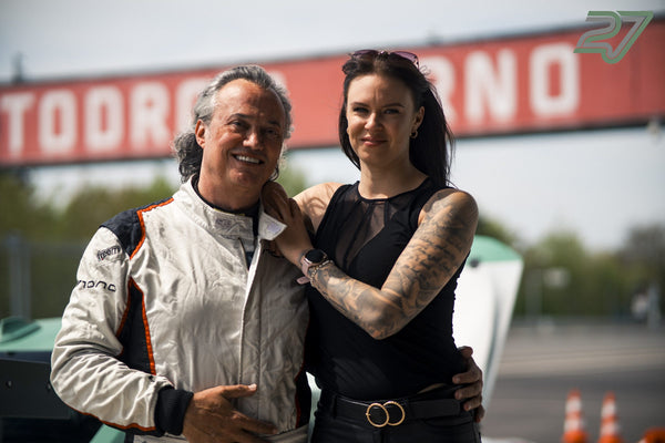 Ein Mann im Rennanzug und eine Frau posieren lächelnd nebeneinander vor dem Schild Automotodrom Brno
