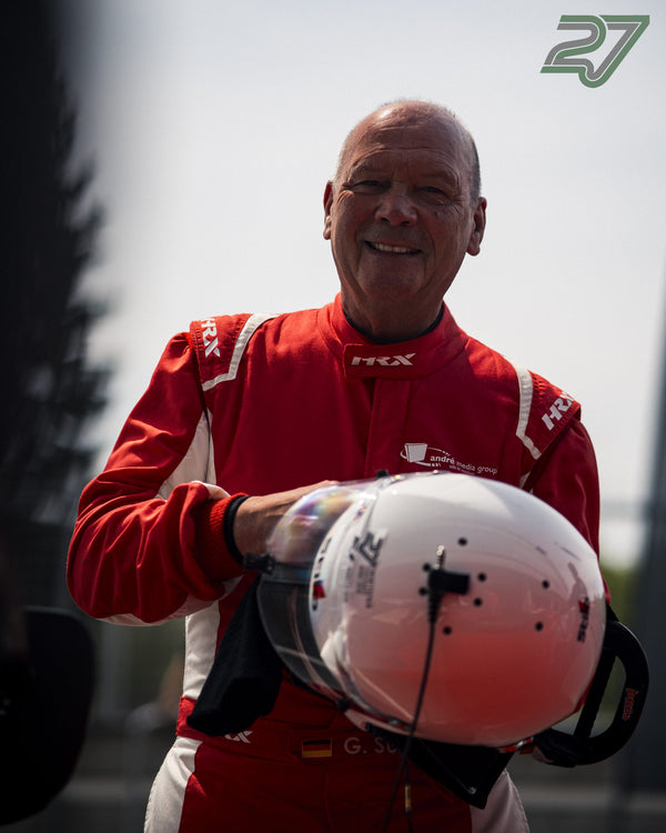 Ein lächelnder Mann im roten Rennanzug mit Sponsorenlogos hält einen weißen Helm unter dem Arm