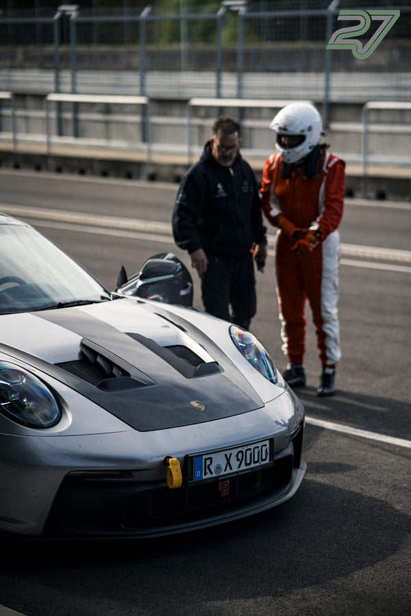 Ein Mann im Gespräch mit einer Person im weiß-roten Rennanzug und Helm neben silbernem Porsche 911 GT3