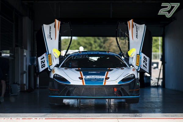 Weiß-blauer McLaren GT3 mit geöffneten Flügeltüren steht in der abgedunkelten Box im Gegenlicht