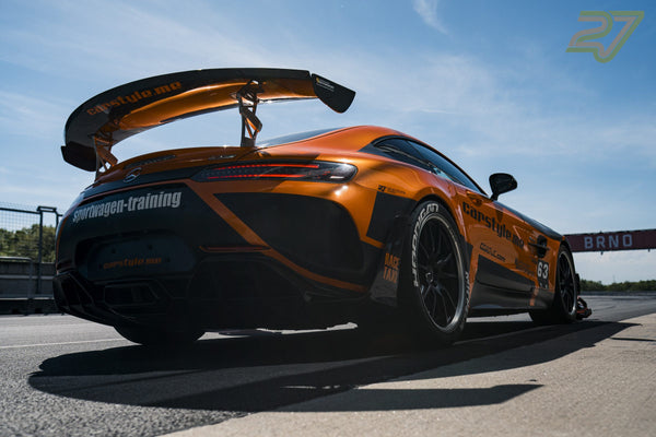 Orange-schwarzer Mercedes-AMG GT rollt heckseitig am BRNO-Schild vorbei, Heckflügel im Gegenlicht