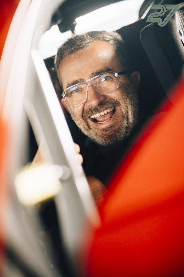 Lächelnder Mann mit Brille lacht herzhaft, durch weiße Überrollbügel und rote Karosserie fotografiert