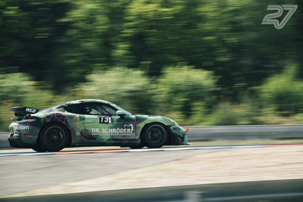 Grün-camouflierter Porsche Cayman GT4 zieht scharf im Seitenprofil an dichtem Grün vorbei