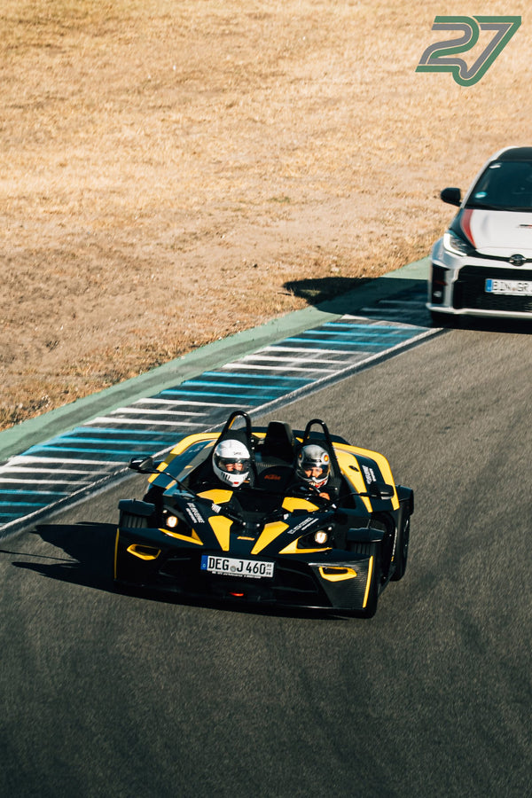 Schwarz-gelber KTM X-Bow mit zwei Helmträgern kurvt vor einem folgenden weißen Rennwagen