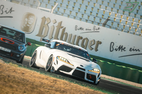 Weißer Toyota Supra mit Rennstreifen jagt an der Bitburger-Bande vorbei, dahinter ein dunkler Mini