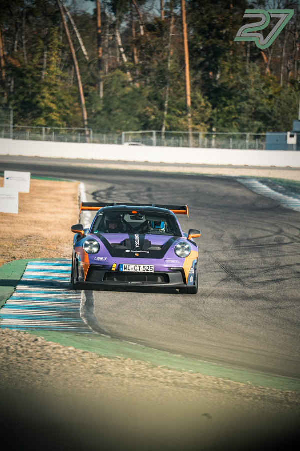 Violetter Porsche 911 GT3 mit Sponsoren-Aufklebern und großem Heckflügel fährt frontal durch eine Kurve