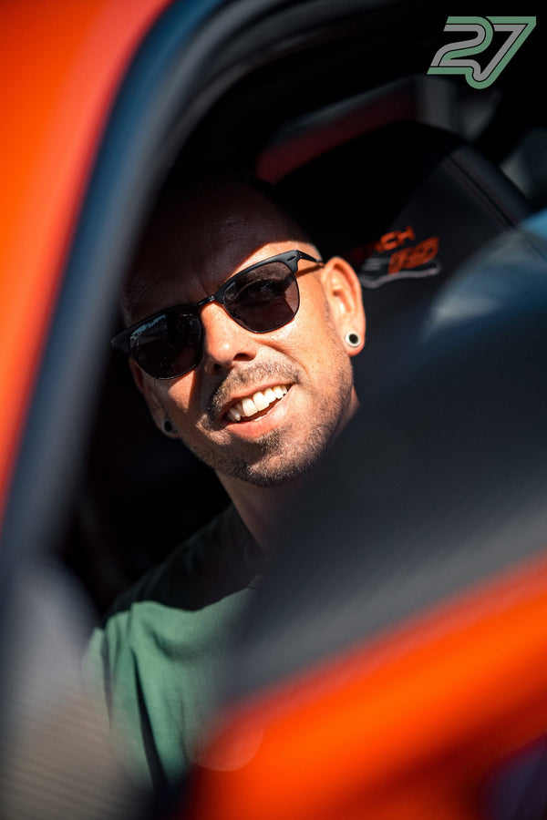 Ein Mann lacht herzhaft durchs Fenster eines orangefarbenen Rennwagens im Fahrerlager