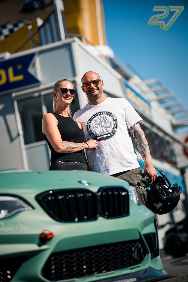 Zwei Teilnehmer posieren lächelnd hinter einem mintgrünen BMW vor dem Boxengebäude