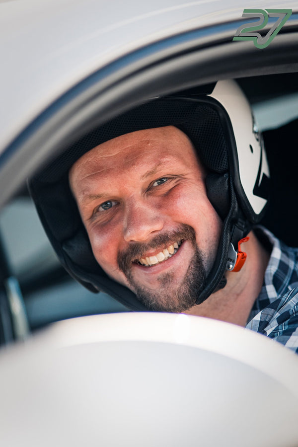 Ein lachender Fahrer mit dunklem Helm sitzt im Auto, kariertes Hemd blitzt hervor