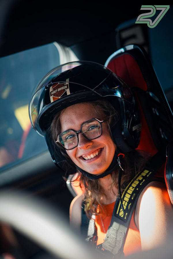 Eine Fahrerin mit Brille und Helm lächelt breit im Cockpit, das Gurtzeug sitzt stramm