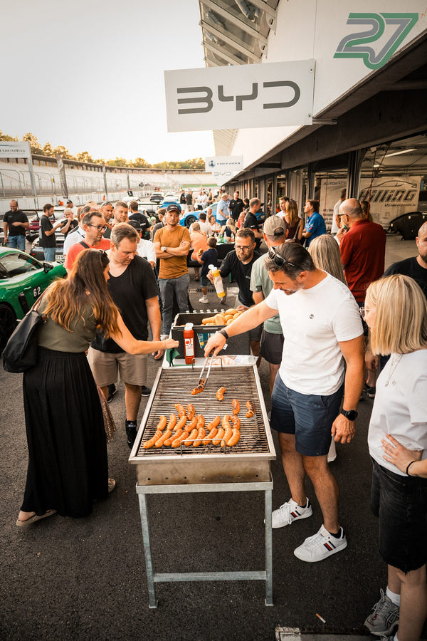 Gästegruppe versammelt sich am Grill, ein Mann wendet Bratwürste vor dem BYD-Schriftzug