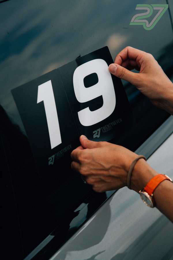 Zwei Hände kleben die schwarze Startnummer 19 auf die Fensterscheibe eines dunklen Autos